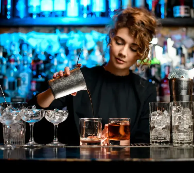 Barman nalewa drink z shakera do szklanki, w tle półki z butelkami i niebieskie światła.