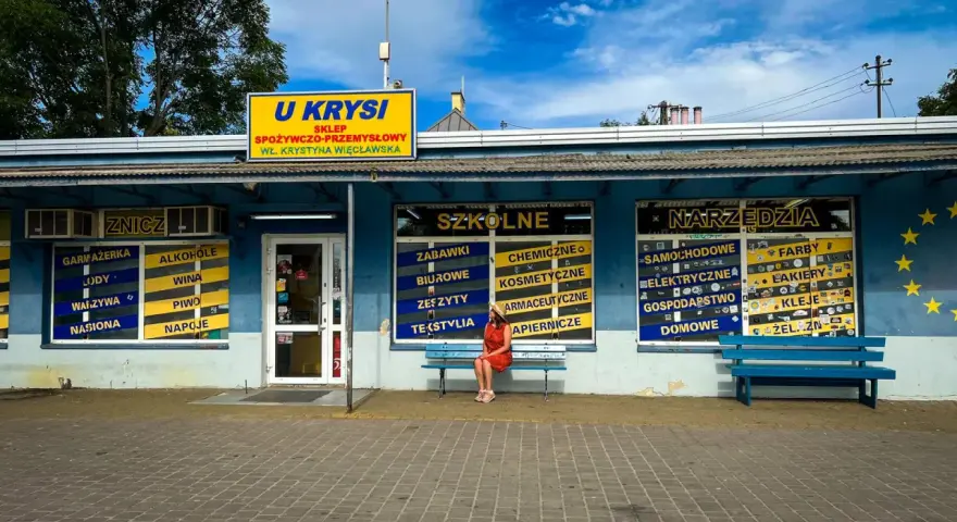 Kobieta w czerwonej sukience siedzi na ławce przed sklepem "U Krysi", na którym widnieją napisy "Szkolne" i "Narzędzia".