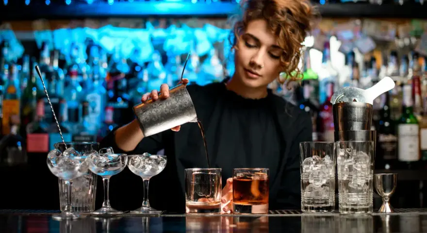 Barman nalewa drink z shakera do szklanki, w tle półki z butelkami i niebieskie światła.